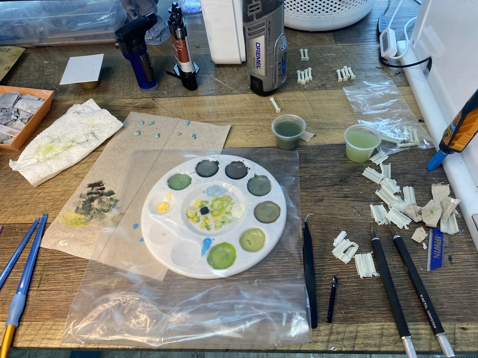 A workbench during model post-processing: paint palette, brushes, hobby knives, a rotary tool, and small 3D-printed parts being painted