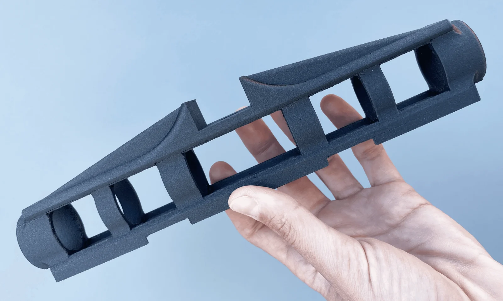 A hand holding a black 3D-printed PA6-CF carbon-fiber nylon part with a matte, grainy surface, against a light blue background