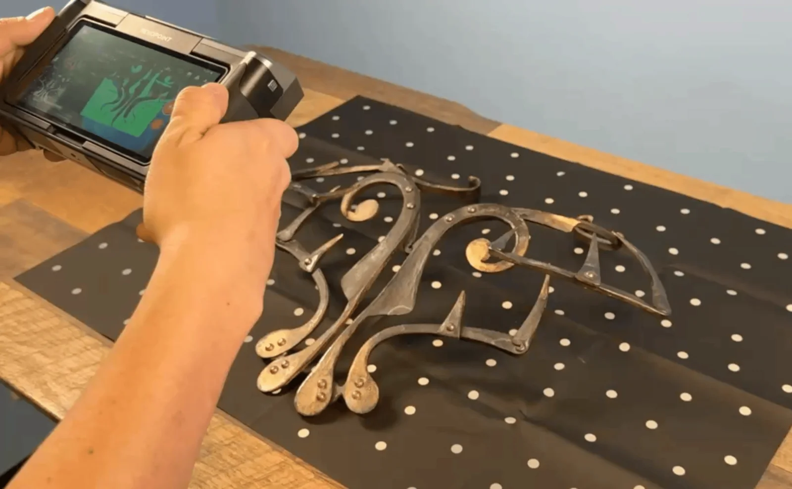 Hands holding a handheld 3D scanner, its screen showing a live capture, while scanning ornate metal hardware laid on a marker-dot mat on a workbench