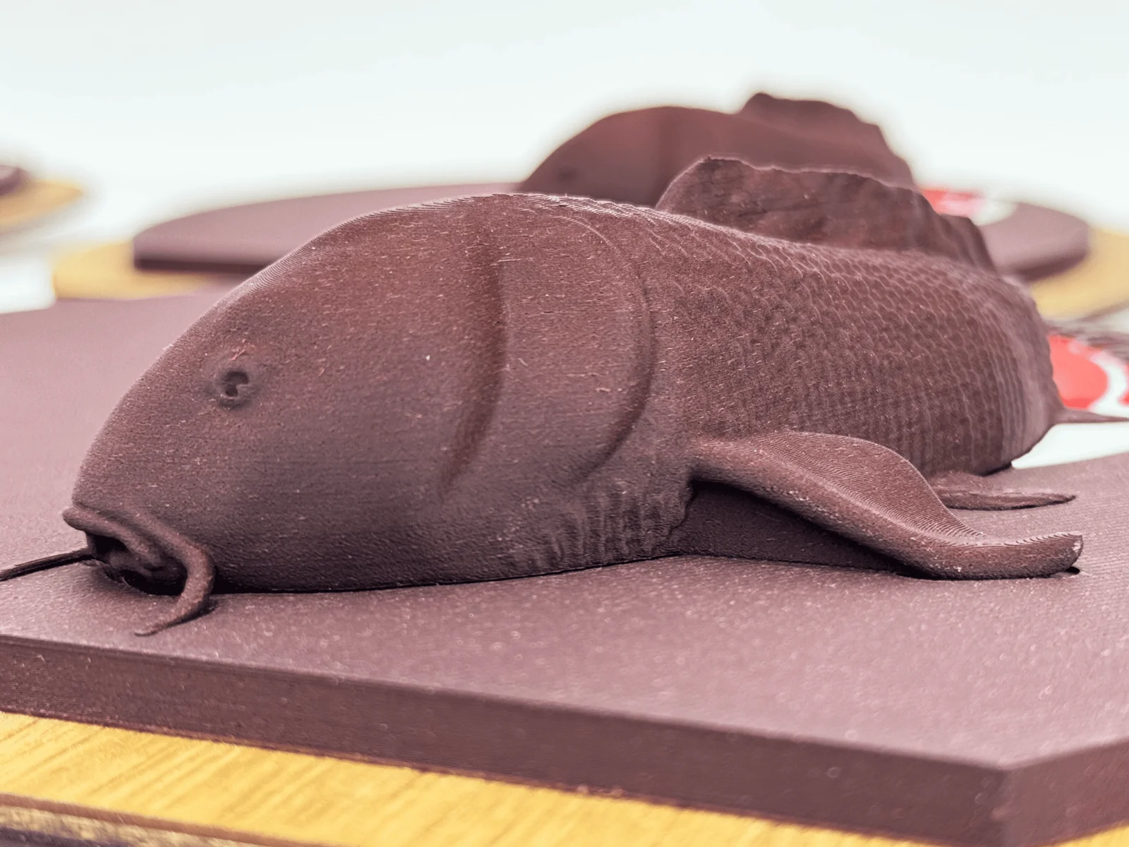 Reddish-brown 3D-printed koi fish sculpture in wood-filled PLA, with scale texture and barbels, resting in half-relief on a matching printed board