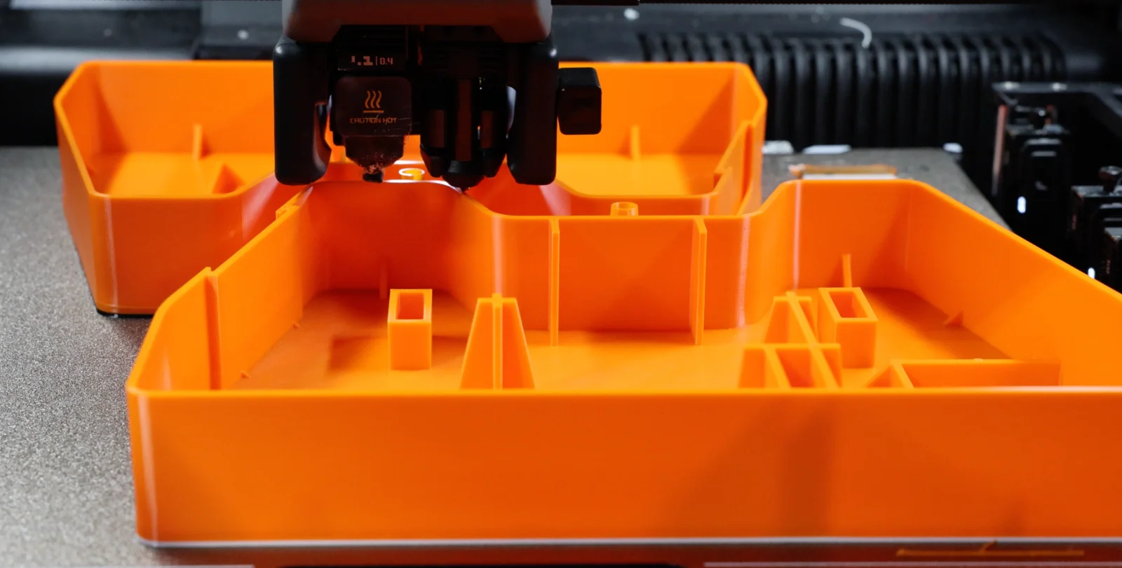 A large orange FDM part being 3D printed, its ribbed internal walls and open bays visible, with the hotend and nozzle at work above it