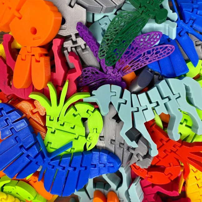 A dense pile of print-in-place articulated toys — axolotls, dragonflies and lizards — in orange, magenta, lime, teal, blue, purple and grey