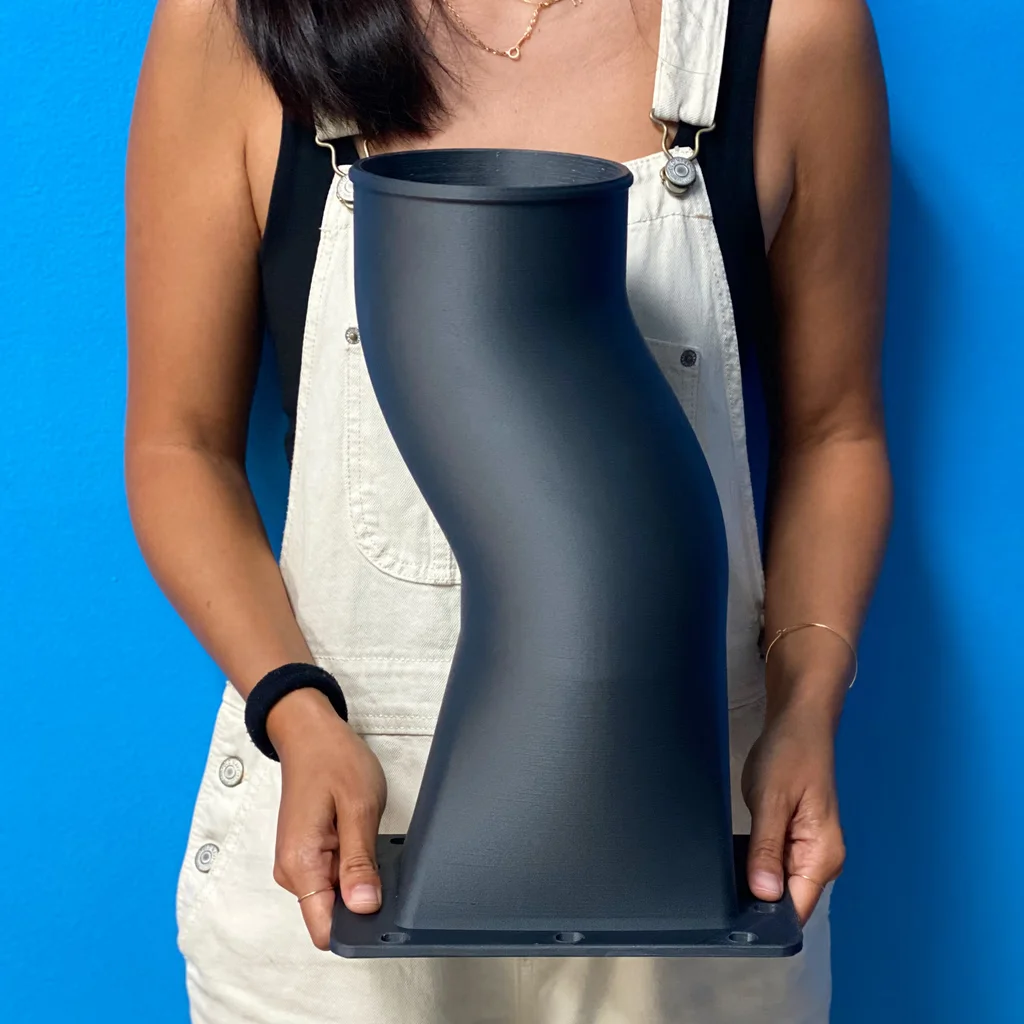 A person holding a large matte black 3D-printed duct transition with a flanged, bolt-holed base